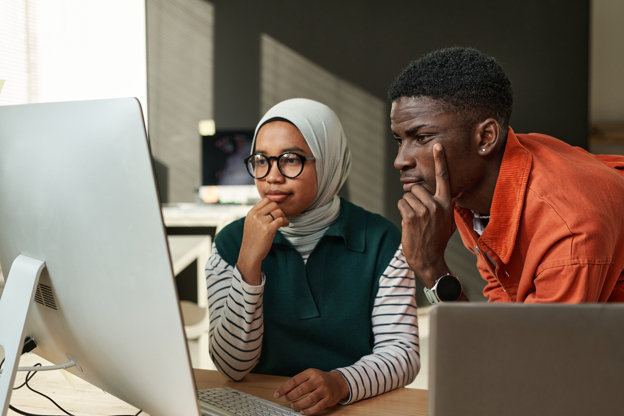 Two young serious intercultural IT engineers looking at data on computer screen or waiting for web page uploading at meeting in office