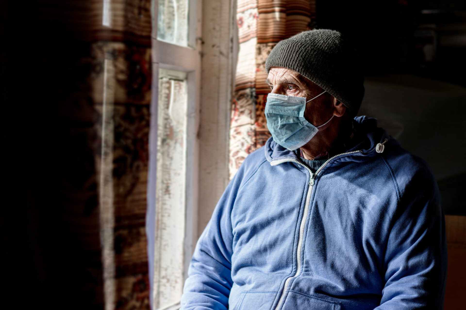 Aged male person wearing facemask and hat looking through window glass indoor